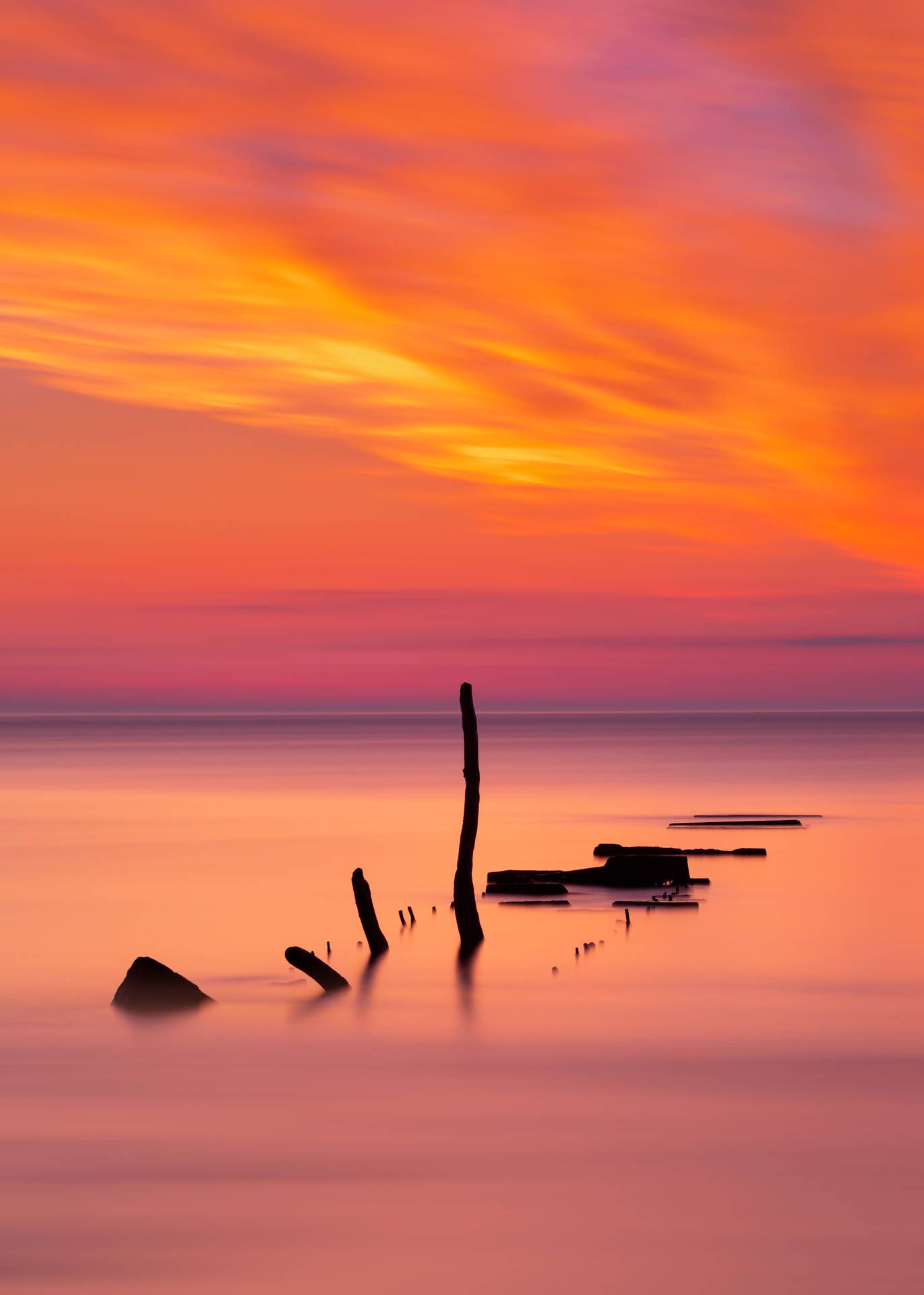 Valtameri oranssilla ja vaaleanpunaisella horisontilla auringonlaskun aikaan, keppejä kellumassa.