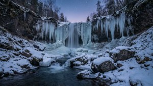 Upea talvinen vesiputous jääpuikkoineen ja lumineen, tähtikirkkaan hämärätaivaan alla.