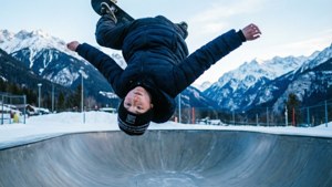 Skateboard-poika tekemässä 360-flippiä puoliputkella lumisessa vuoristomaisemassa.