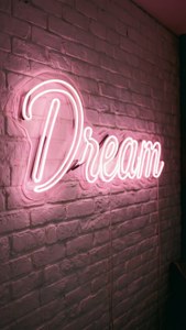 Vaaleanpunainen neonkyltti 'Dream' hehkuu valkoisella tiiliseinällä, inspiroiva ja esteettinen. — Vaaleanpunainen esteettinen Taustakuvat